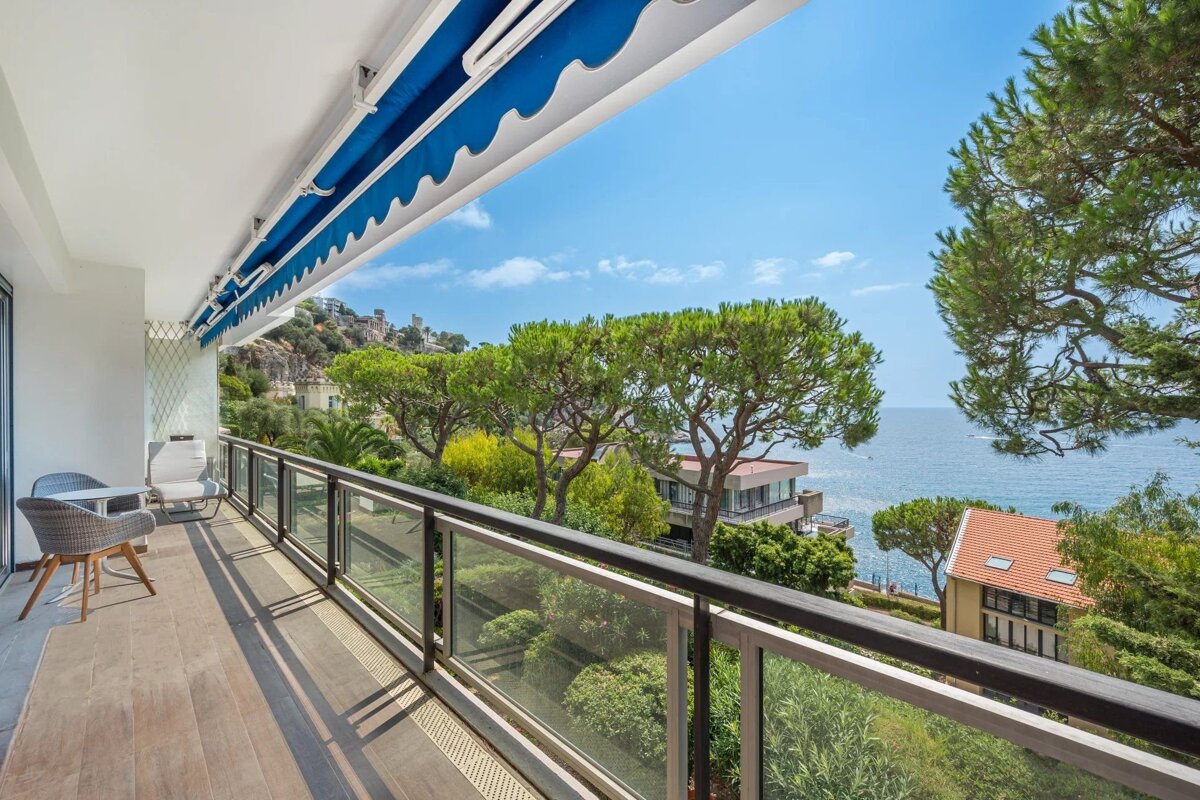 A sunny balcony with a blue awning overlooks lush green trees, a small town, and the blue ocean under a clear sky.