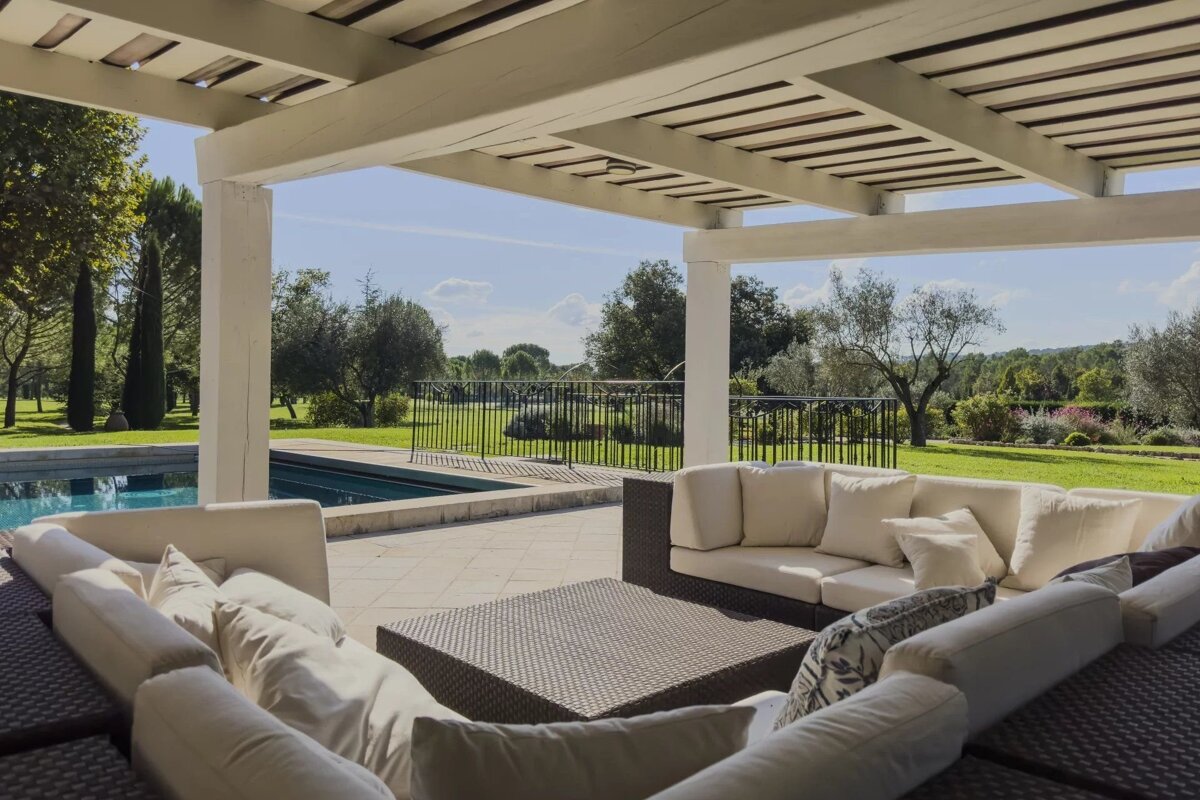 Luxurious outdoor lounge under a white pergola, featuring comfortable sectional sofas next to a swimming pool and scenic lawn.