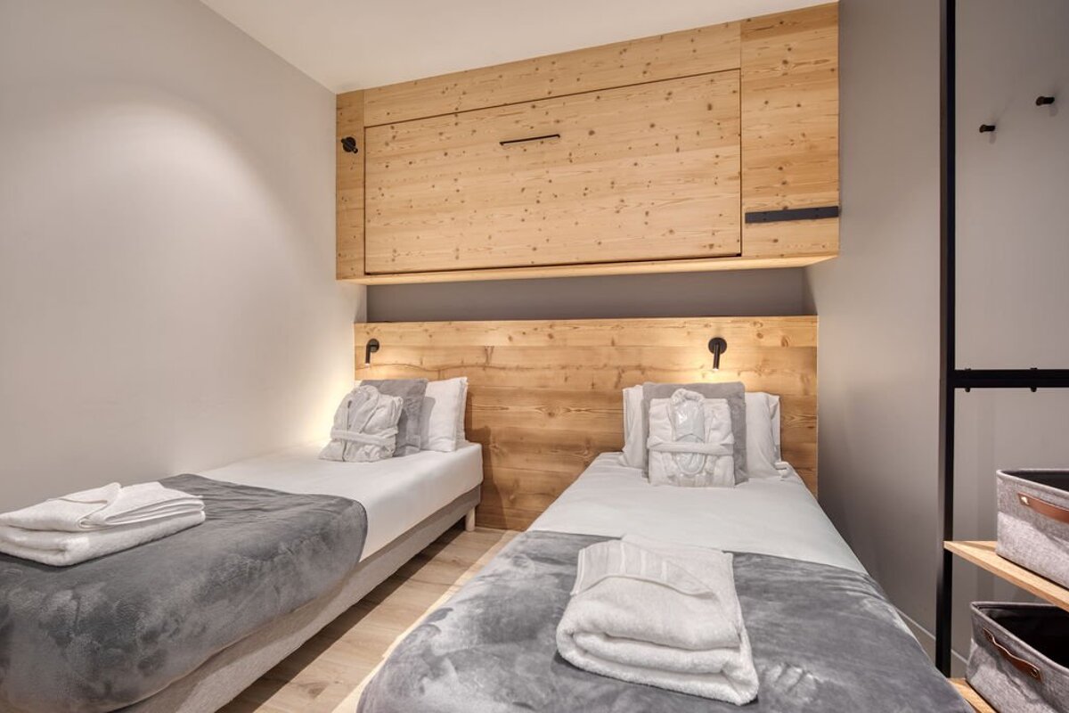 A modern bedroom features two single beds with white and grey bedding, bathrobes, and light wood paneling with an integrated wall unit.