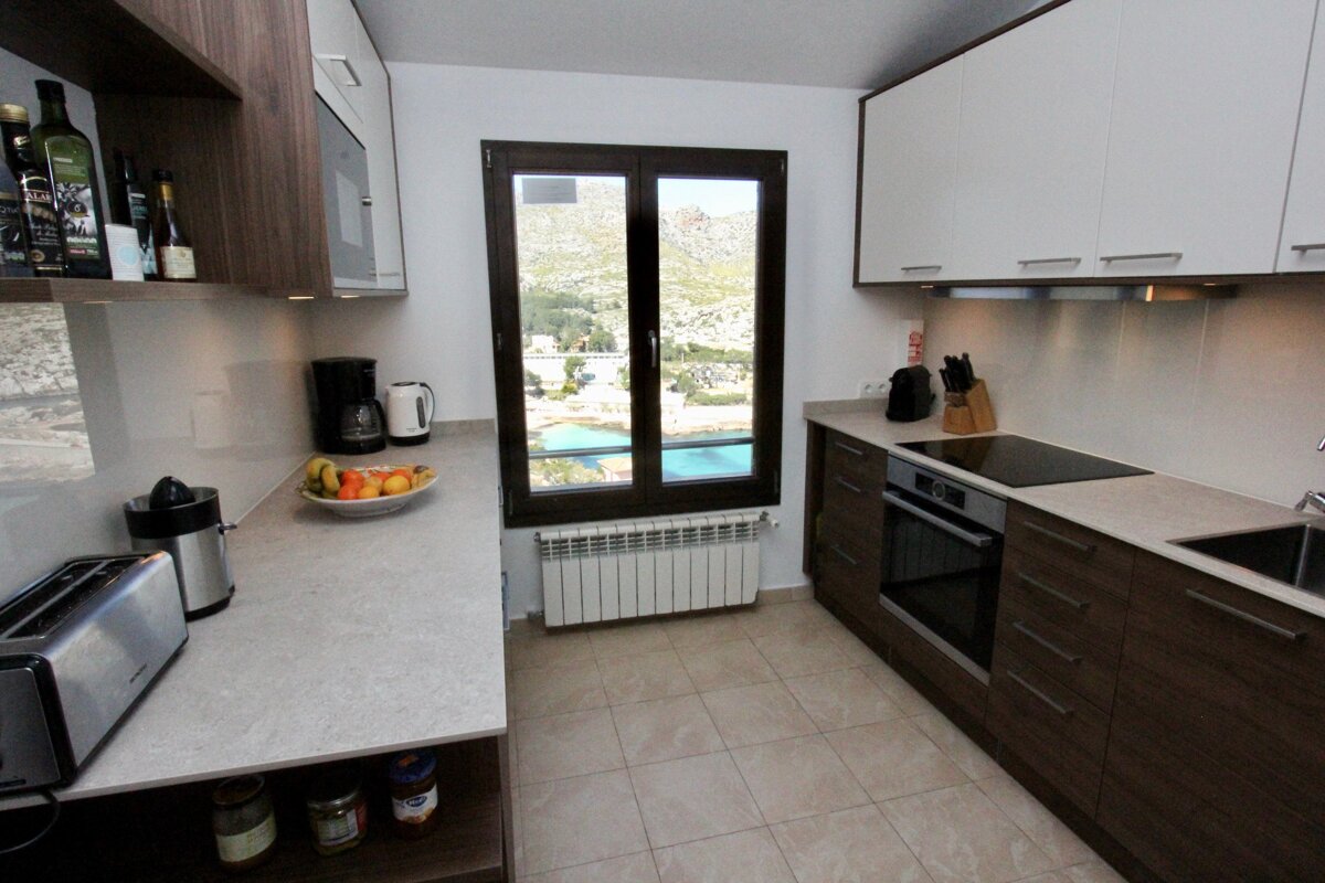 A modern kitchen with brown and white cabinets, a window overlooking mountains and turquoise water, and appliances like a toaster and coffee maker.