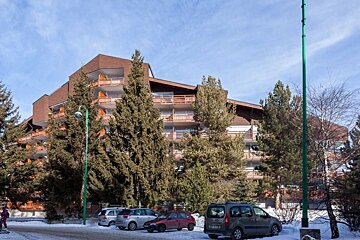 Multi-story resort building with balconies, surrounded by pine trees and parked cars on a snowy day.