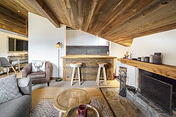 Cozy chalet interior with slanted wooden ceilings, a rustic fireplace, living area, and a kitchen bar counter with wooden stools.