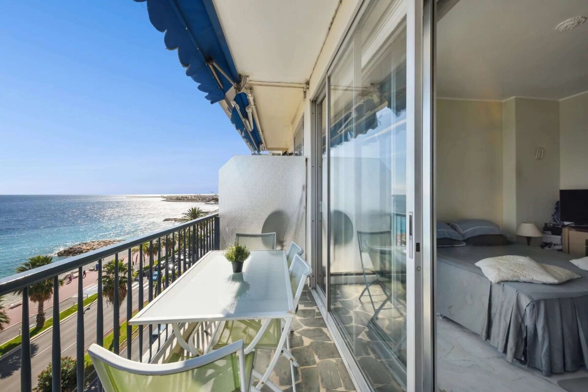 Sunny balcony with dining table and chairs, offering expansive ocean and coastal road views. Sliding doors lead to a bedroom, with a blue awning overhead.