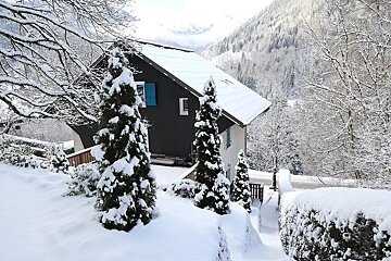 A dark house and evergreen trees are blanketed in fresh snow, with snowy mountains and forests in the background, creating a winter scene.