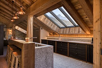 Cozy, rustic kitchen in a wooden chalet. Exposed beams, copper lights, and large skylights frame stunning snowy mountain views. Features a granite-topped bar.