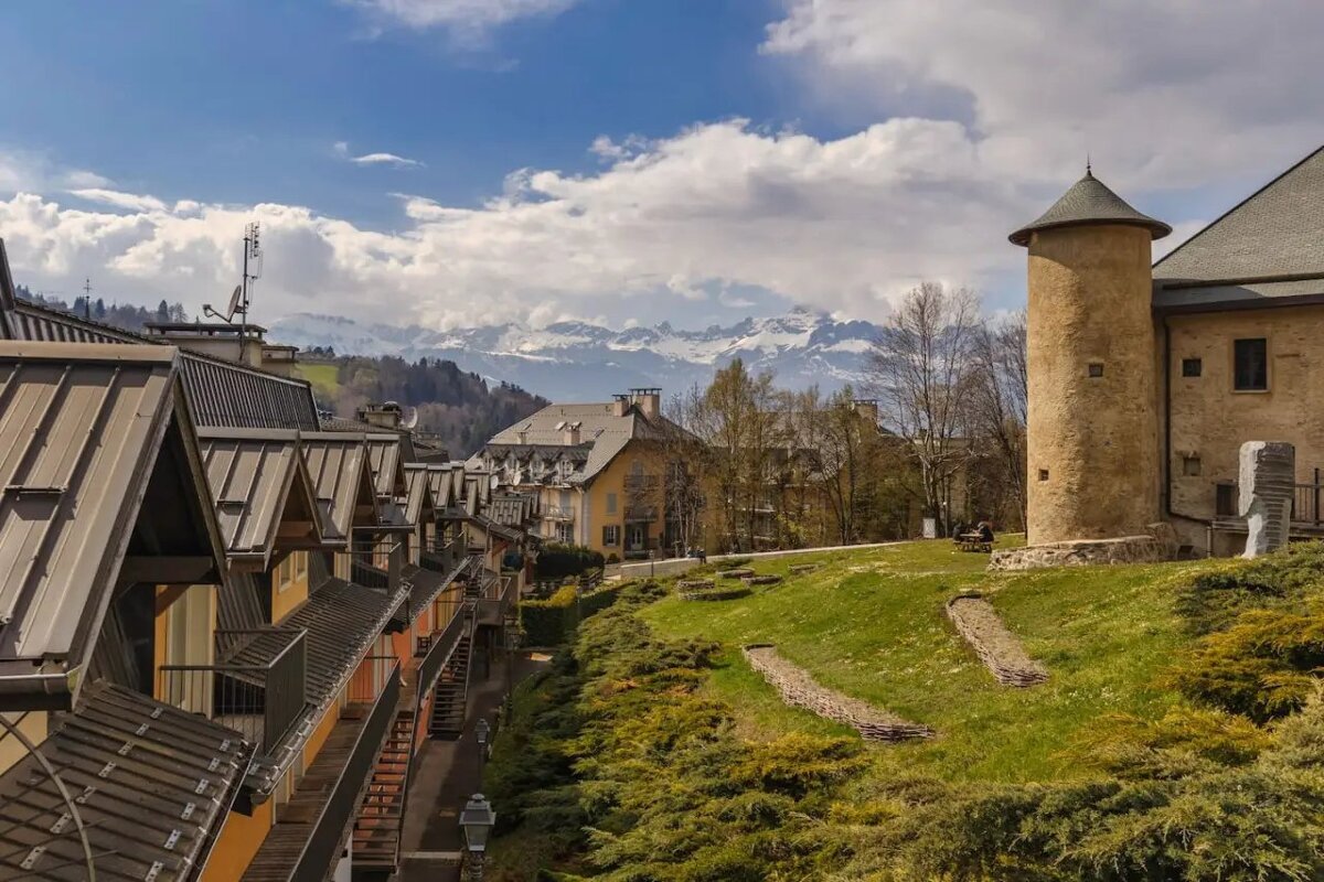 Picturesque alpine village with traditional houses, a stone tower on a grassy hill, and snow-capped mountains in the distance.