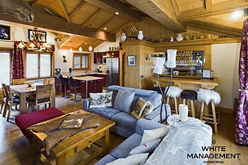 A rustic chalet-style open-plan living room, dining area, kitchen with red cabinets, and bar with furry stools, all under a beamed wooden ceiling.