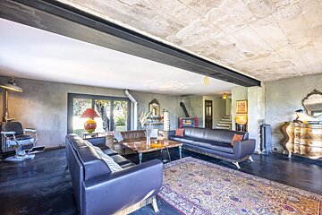 An eclectic living room features leather sofas, a barber chair, vintage furnishings, concrete walls/ceiling, and dark floors with a view outside.