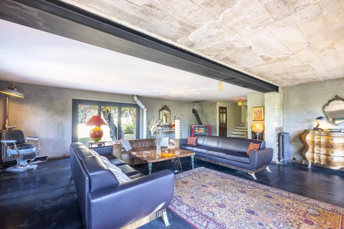 Industrial-style living room with exposed concrete ceiling, leather sofas, a vintage barber chair, and a patterned rug on dark floors.