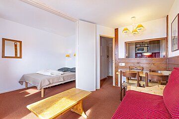 A compact apartment with a bed in an alcove, a living/dining area with a red sofa, and a kitchen visible through a pass-through. Features wood paneling and red carpet.