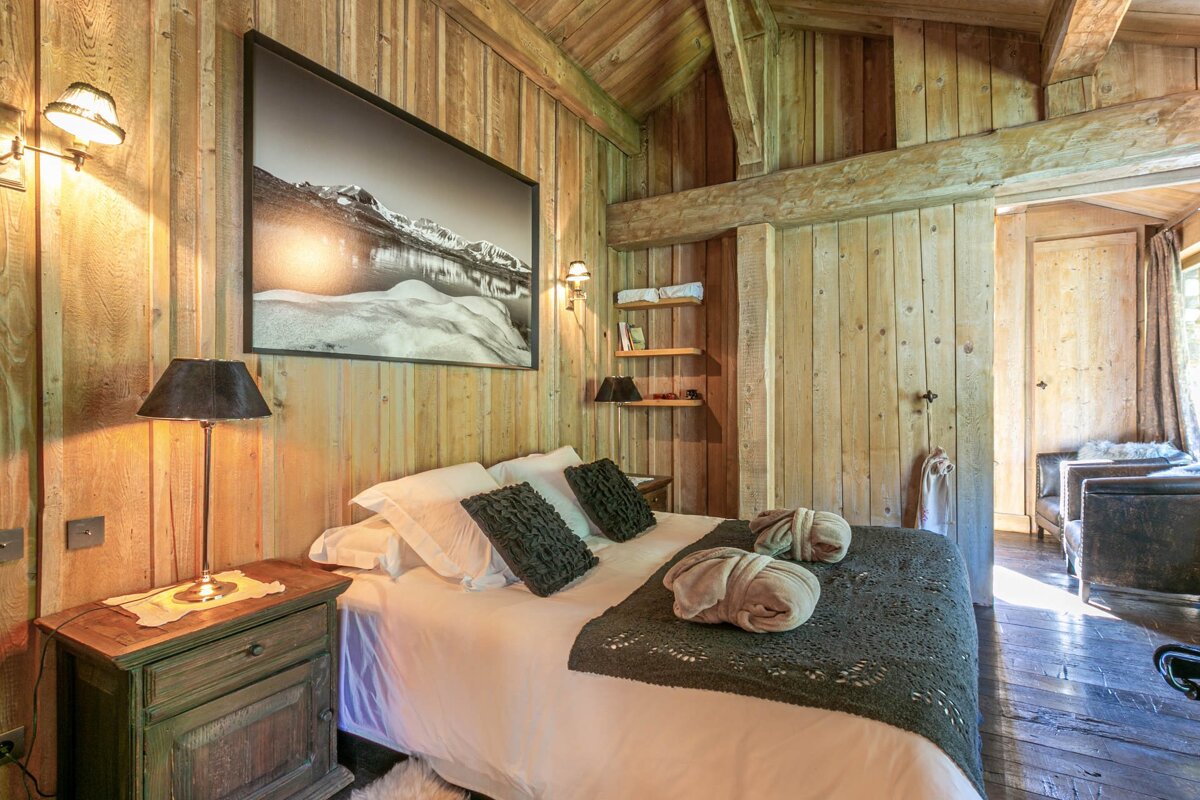 A cozy, rustic bedroom with wood-paneled walls, ceiling, and exposed beams. A large bed with dark bedding and a mountain artwork dominates the space, creating a warm, cabin-like atmosphere.