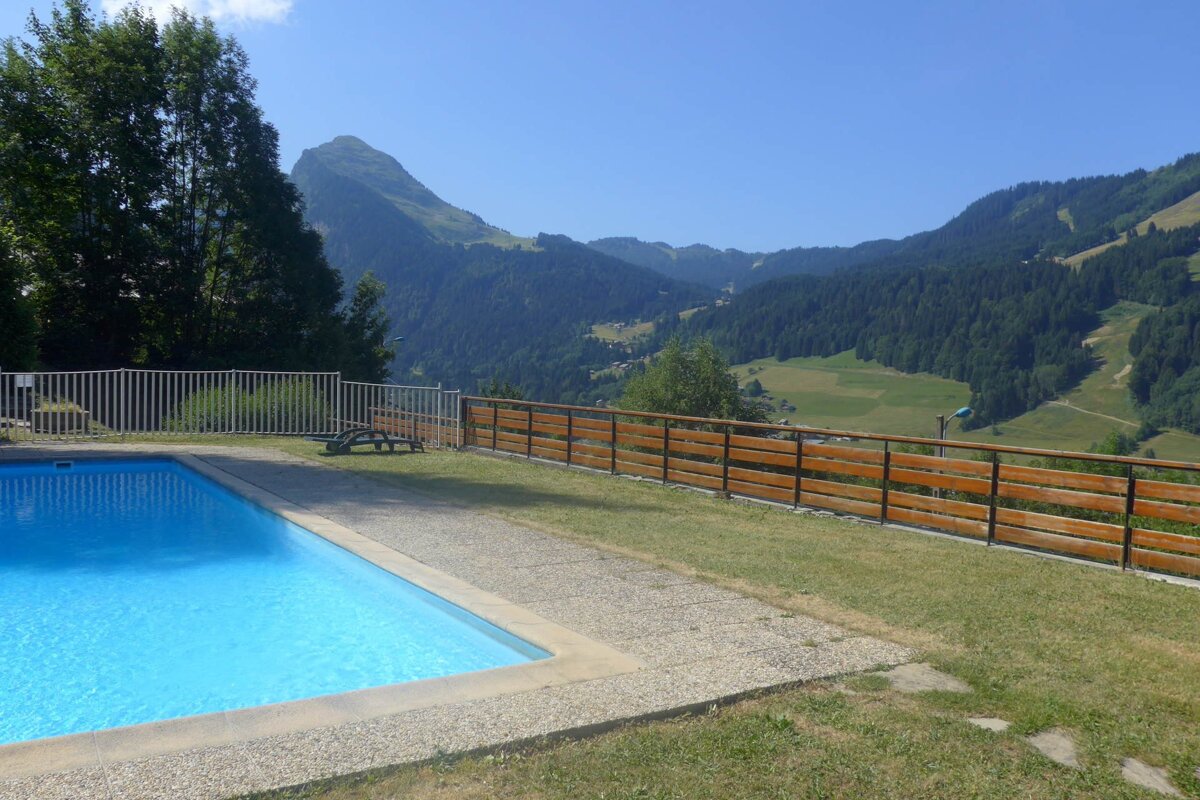A bright blue swimming pool overlooks majestic green mountains and a lush valley under a clear, sunny sky, with a wooden fence along the hillside.