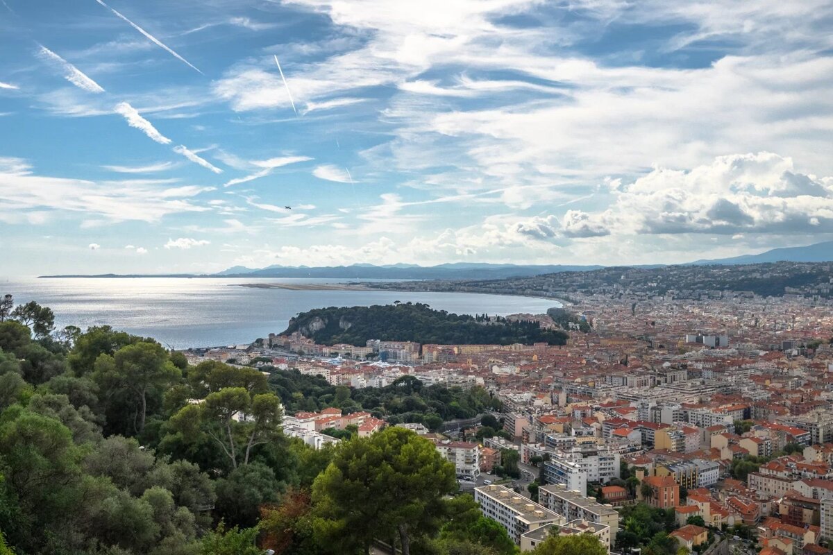 High-angle view of a coastal city with a bay, green hills, and a partly cloudy sky.