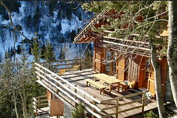 A wooden house with a large deck surrounded by snow covered trees
