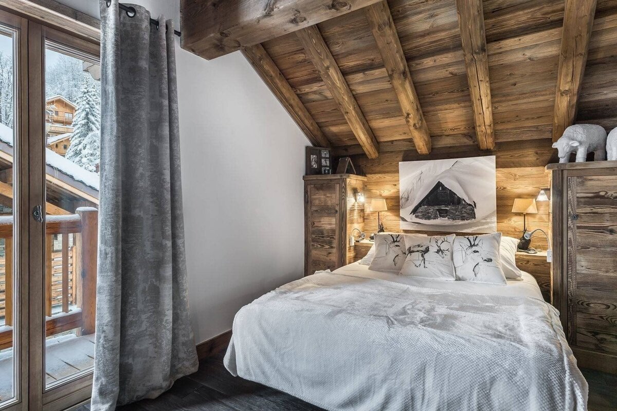 Cozy, rustic chalet bedroom with wooden walls, beamed ceiling, a large bed, and a balcony overlooking a snowy winter landscape.