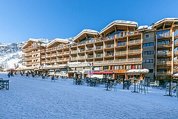 Sunny ski resort scene with modern wooden apartment buildings, outdoor cafe seating on a snowy piste, and distant mountains.