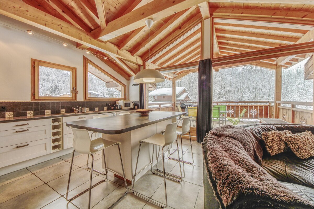 Cozy chalet interior featuring a modern kitchen island, comfy sofa, rustic wood beams, and large windows revealing a beautiful snowy forest view.