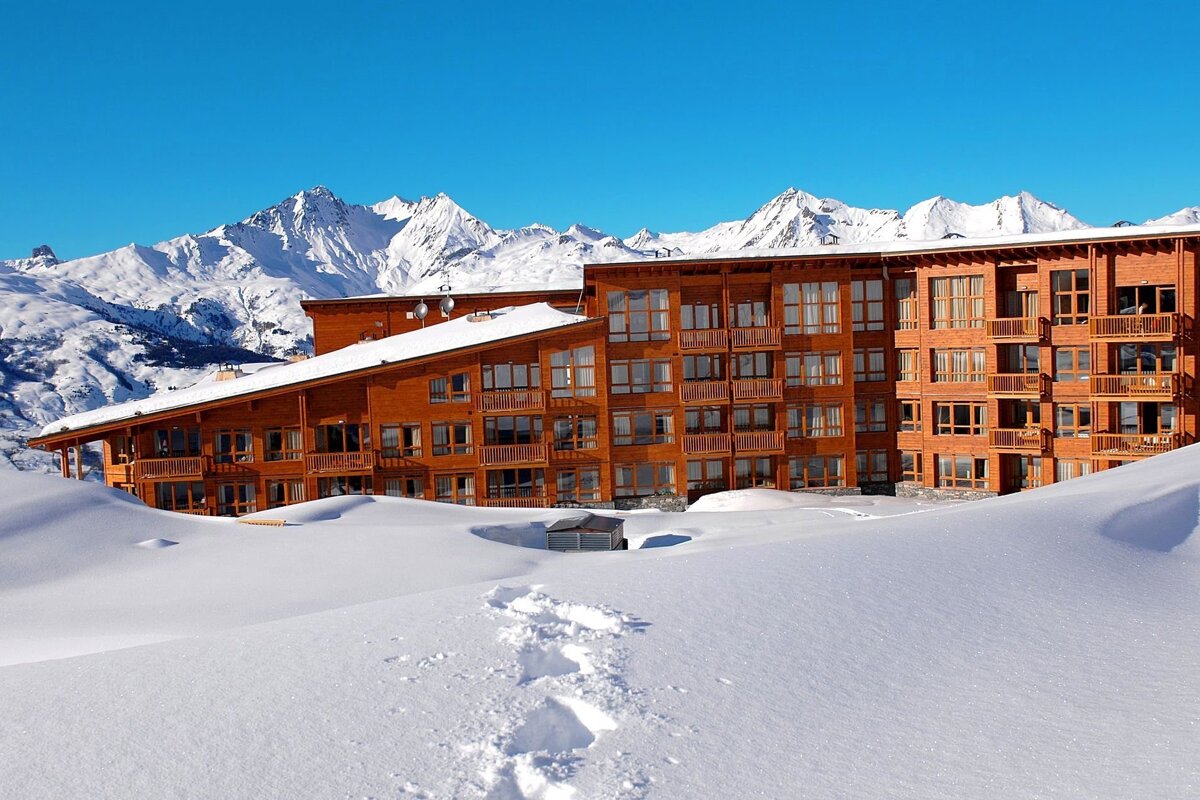 A large timber building sits in deep snow with footprints leading towards it, backed by majestic snow-capped mountains under a clear blue sky.