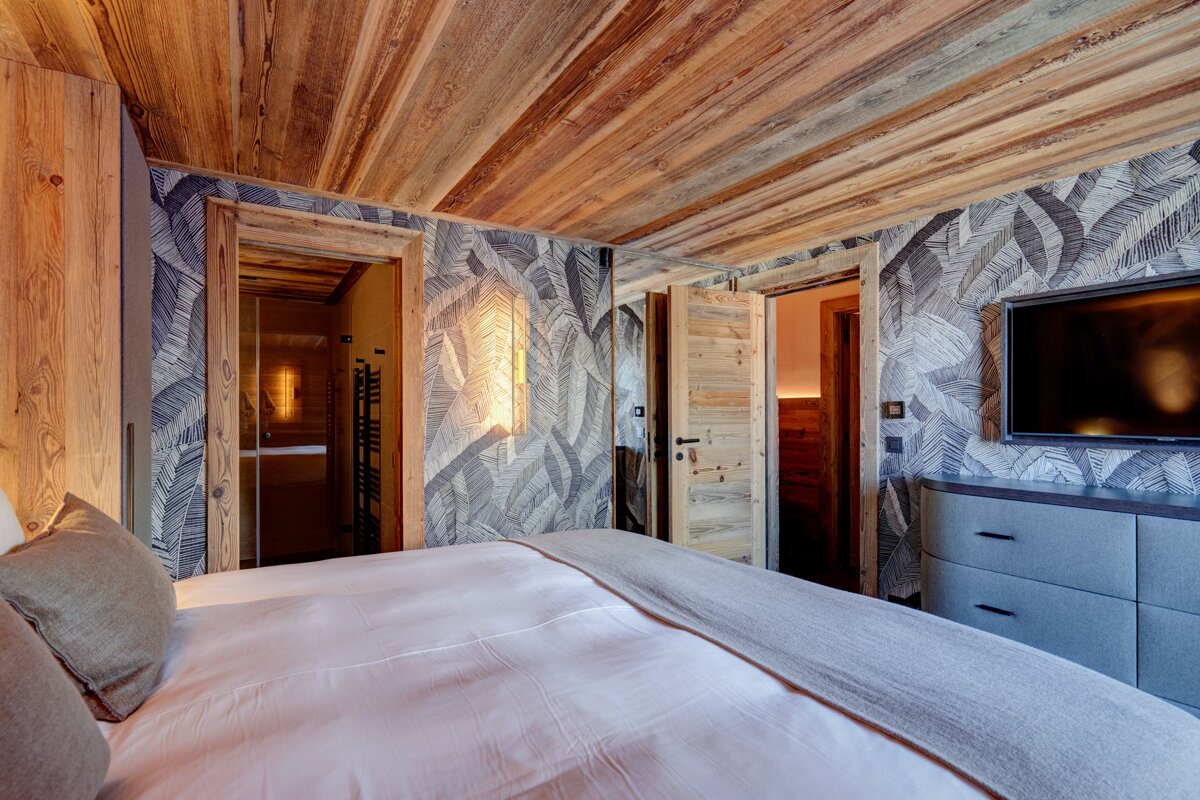 A cozy bedroom with a rustic wooden ceiling, patterned leaf wallpaper, a bed, and wooden doors leading to a bathroom and another room.