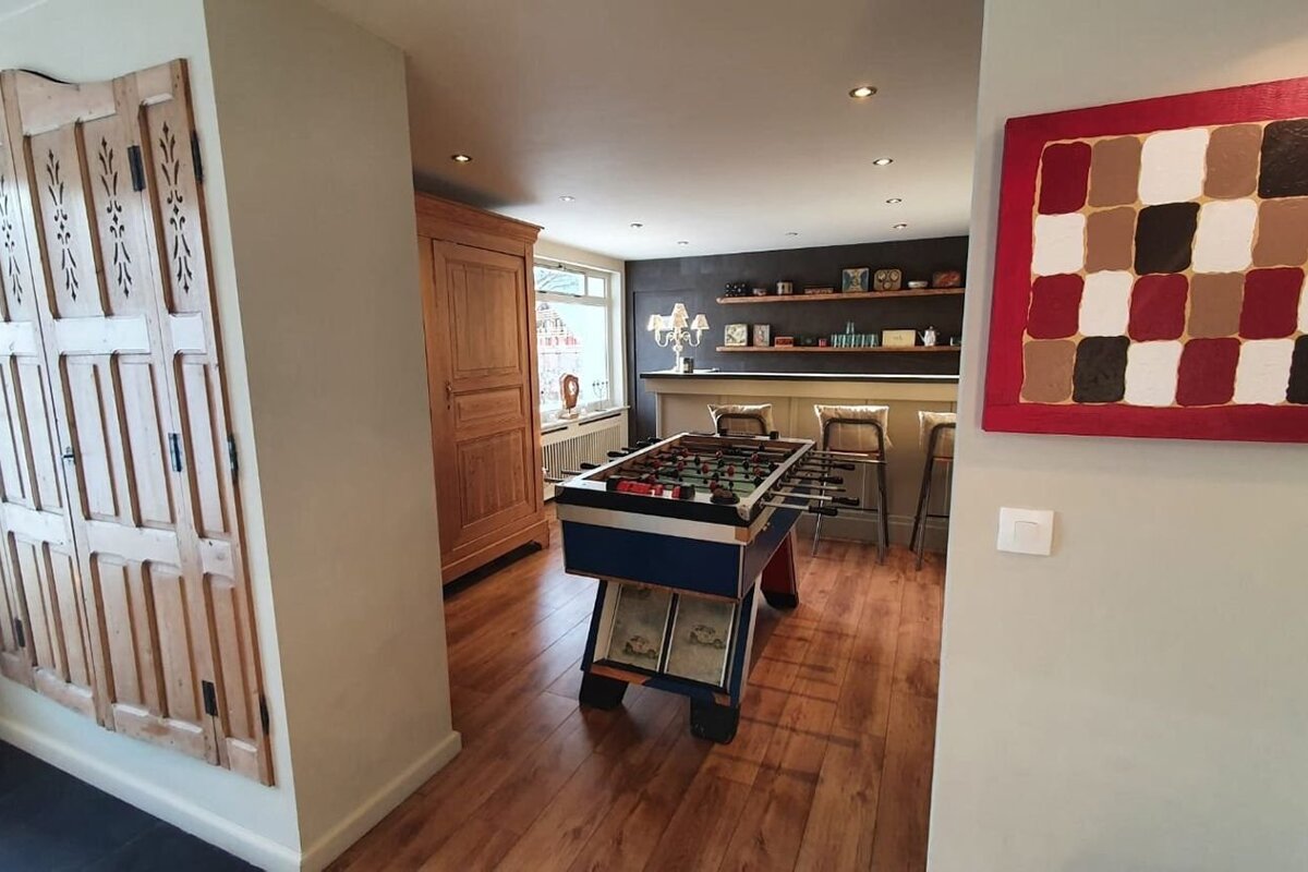 A well-lit recreation room featuring a foosball table, a bar with stools, wooden floors, a large wooden cabinet, and abstract wall art.
