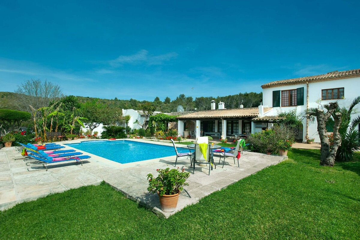 A picturesque white villa with a sparkling blue pool, surrounded by a vibrant green lawn and abundant foliage under a clear sky. Lounge chairs await.