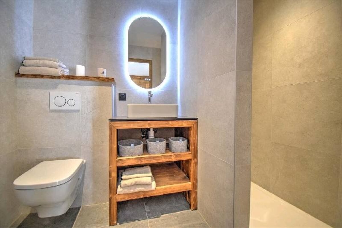 Modern bathroom with a backlit oval mirror, wooden vanity, vessel sink, wall-mounted toilet, and grey tiles.