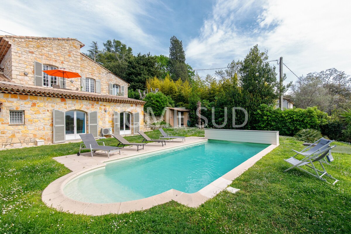 A large swimming pool is surrounded by chairs and umbrellas in front of a house that is for sale