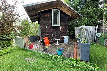 A small, rustic wooden cabin with a deck featuring colorful planters and modern chairs, surrounded by lush green grass and trees.