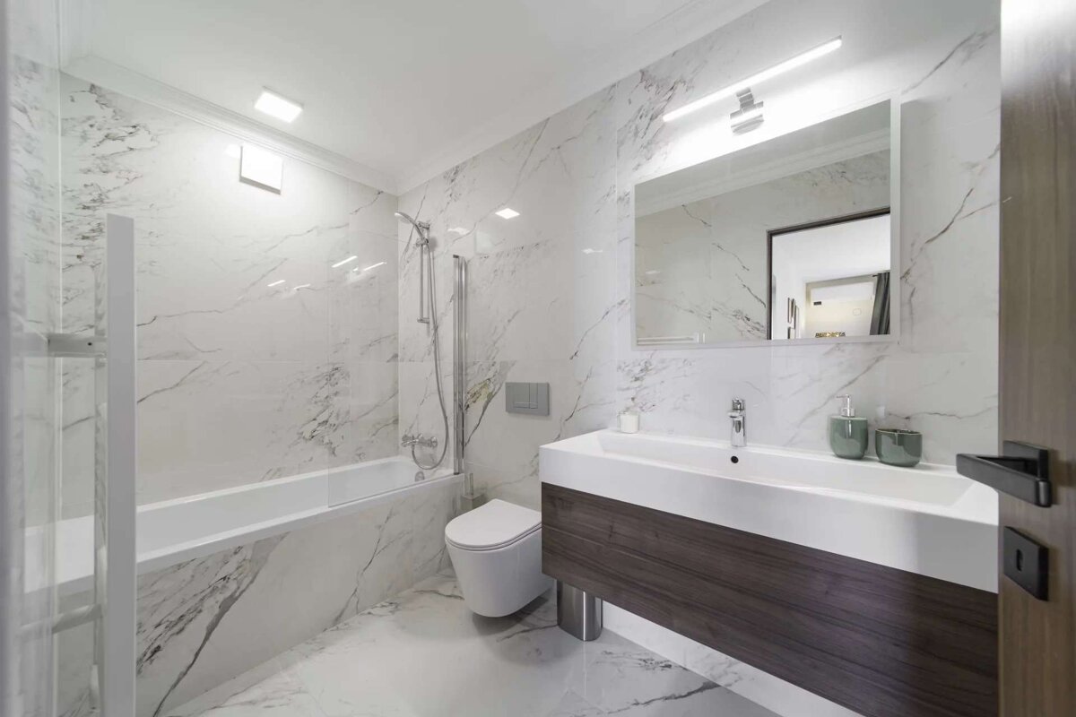 A bright, modern bathroom showcasing white marble walls and floor. It features a bathtub with shower, toilet, and a sleek vanity with a sink.