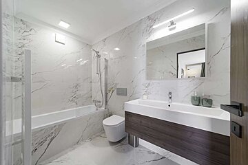 A bright, modern bathroom showcasing white marble walls and floor. It features a bathtub with shower, toilet, and a sleek vanity with a sink.