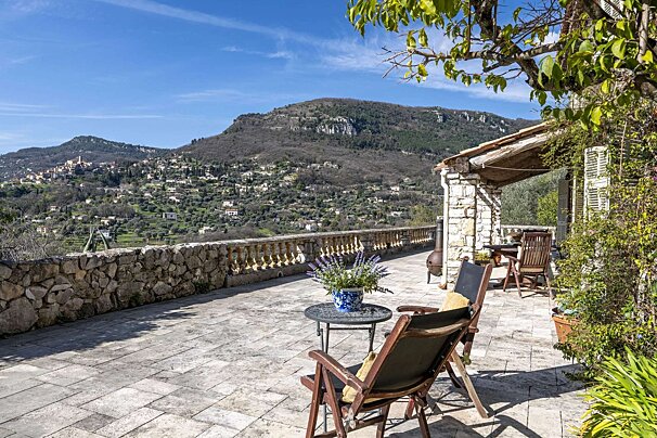 Sunny stone terrace with chairs and lavender, offering panoramic views of a distant village nestled in a lush valley and rugged mountains.