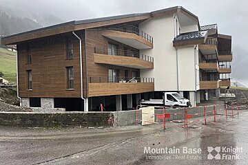A contemporary building with wood siding and white walls, featuring balconies, in a misty mountain landscape. A white truck and construction barriers are visible.
