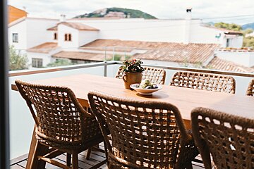 A balcony dining table with wicker chairs and a plant, overlooking white houses with red roofs under a cloudy sky.