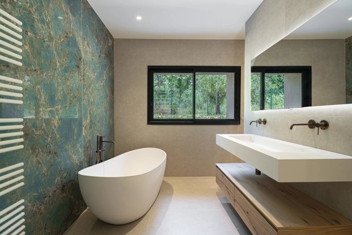 Sleek modern bathroom with a teal marble accent wall, freestanding tub, double vanity, and a window offering a lush green view.