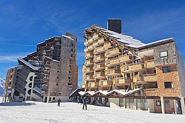 A large, multi-story wooden chalet-style building with balconies, covered in snow, against a backdrop of vast snowy mountains.