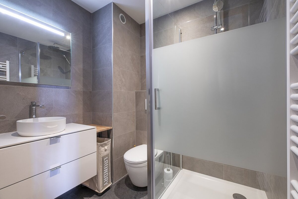 Modern bathroom with grey tiles, a white vanity and vessel sink, toilet, laundry bin, and glass-enclosed shower.