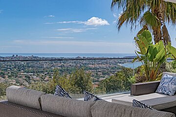 An outdoor balcony with wicker seating and palm trees overlooks a vast coastal city and the ocean under a blue sky.