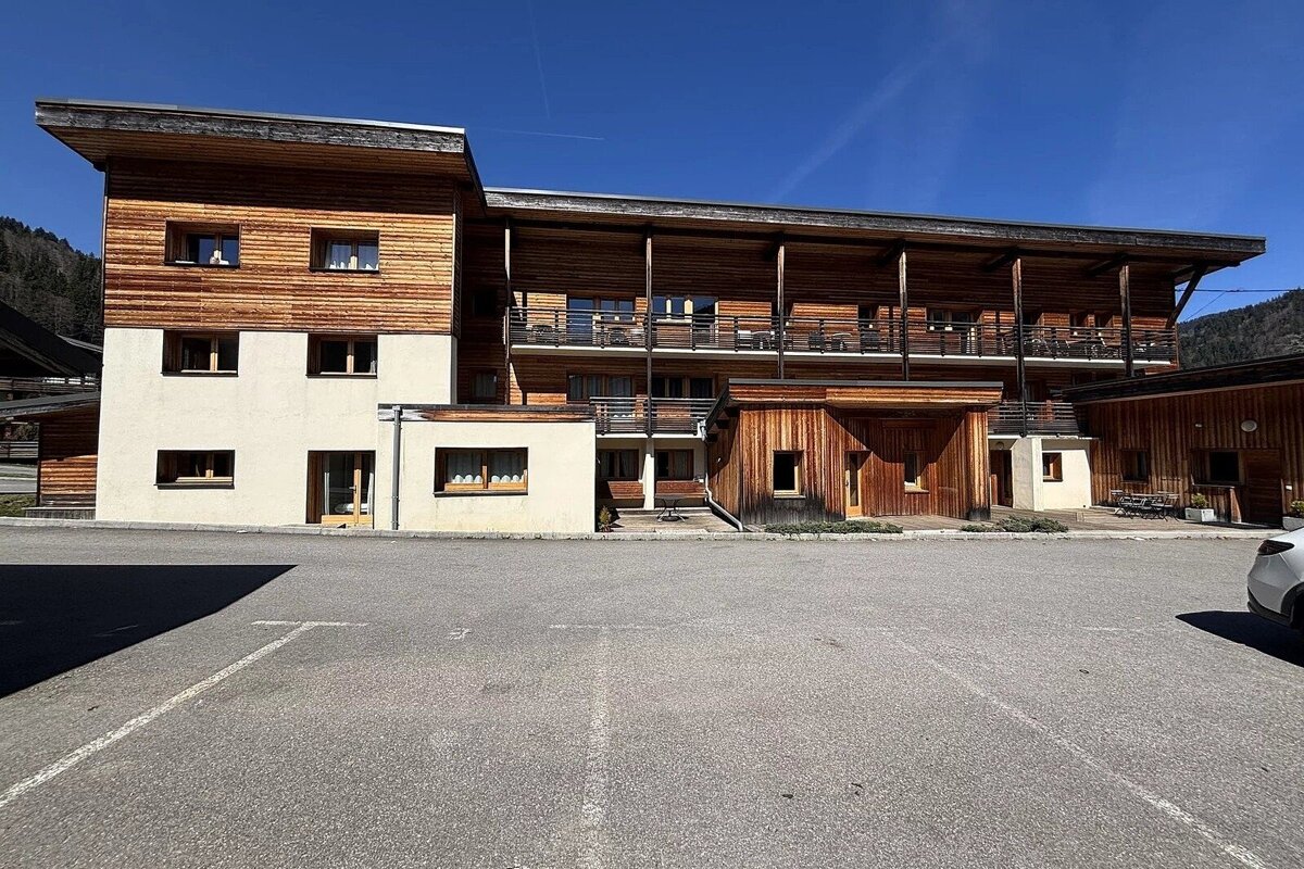 A multi-story modern building with wood and light walls, balconies, and an empty paved parking lot in front, all under a clear blue sky.