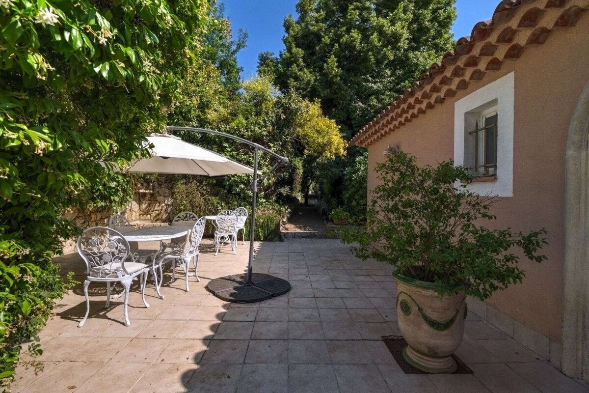 A sunlit outdoor patio with a white metal table and chairs under an umbrella, surrounded by lush greenery, next to a house with a tiled roof.