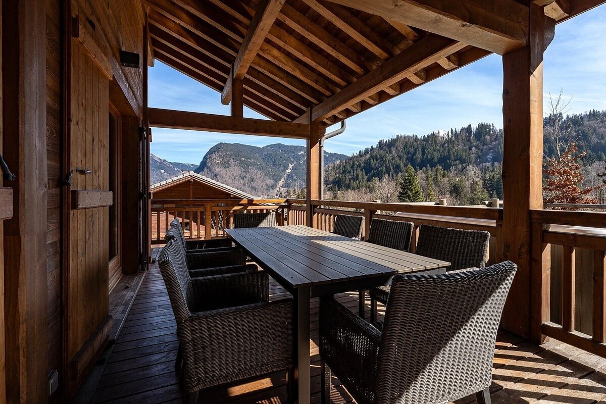 A beautiful wooden terrace with an outdoor dining table and chairs, offering panoramic views of mountains and forests under a clear sky.