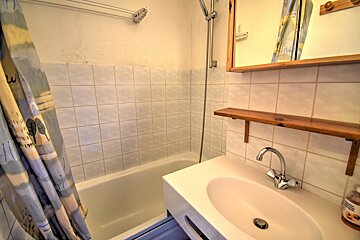 A compact bathroom features a bathtub with a patterned shower curtain, a wall-mounted shower, a white sink, a mirror, and a wooden shelf.