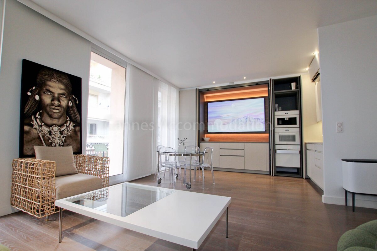 A modern open-plan living room with an integrated kitchen, dining table, large TV, woven armchair, and white coffee table on hardwood floors.