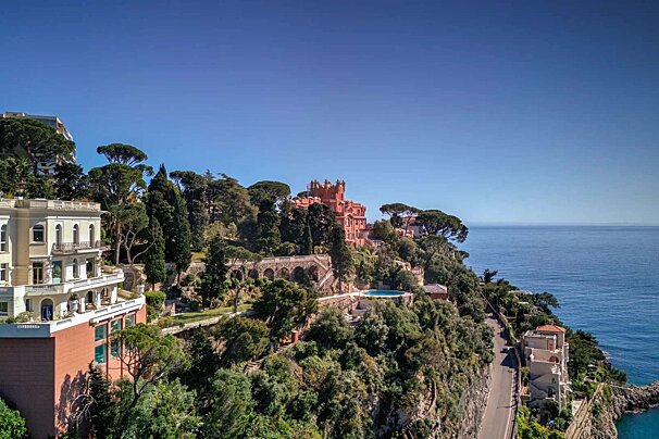 A grand red castle, white villas, and lush greenery cling to a steep cliff overlooking the blue sea, with a coastal road curving along.