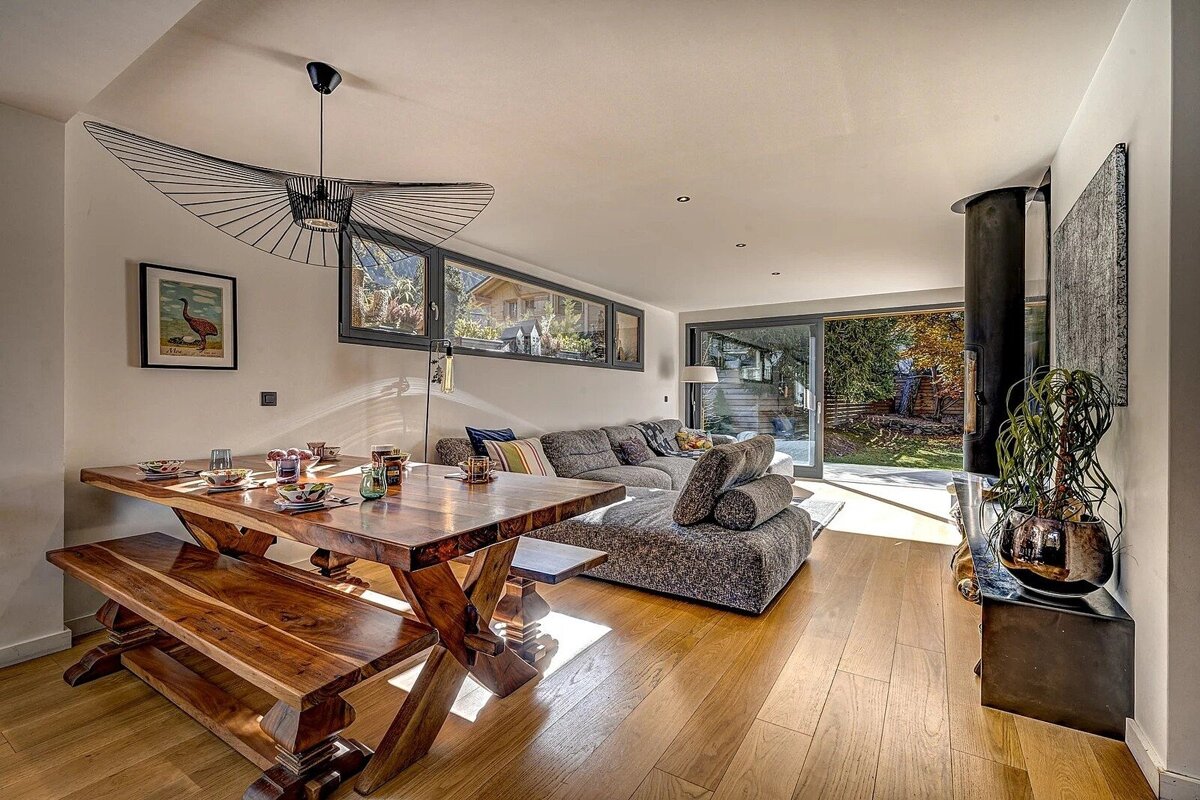 Bright, open-concept living space with a rustic wooden dining table, modern chandelier, comfy grey sectional sofa, and large windows offering garden views.