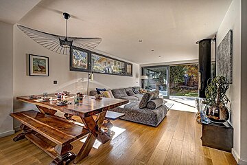 Bright, open-concept living space with a rustic wooden dining table, modern chandelier, comfy grey sectional sofa, and large windows offering garden views.