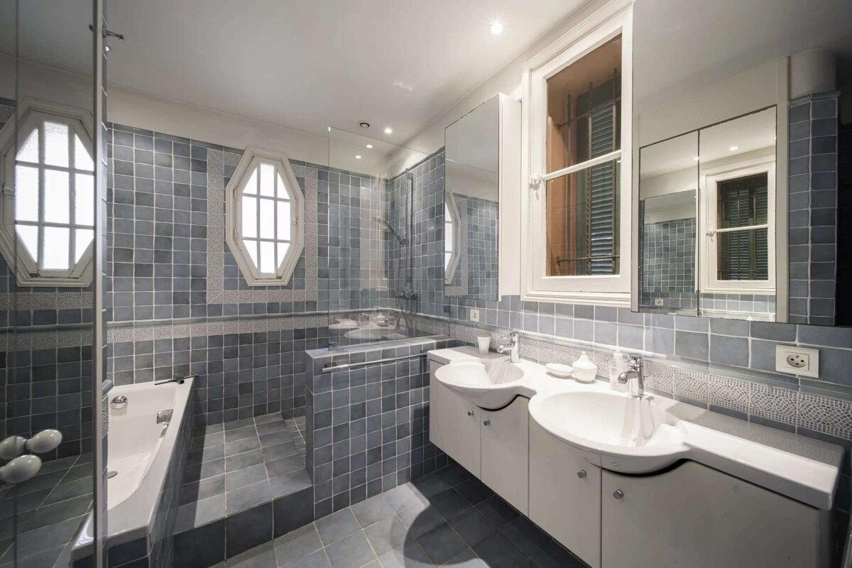 A modern blue-tiled bathroom featuring a bathtub, walk-in shower, double vanity, and unique octagonal & rectangular windows.