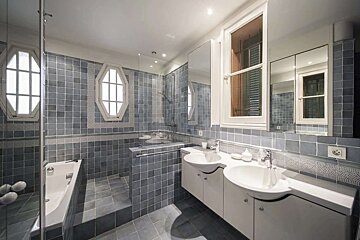 A spacious bathroom with blue-grey tiles, featuring a bathtub, shower, dual sinks, and multiple windows, including unique octagonal ones.