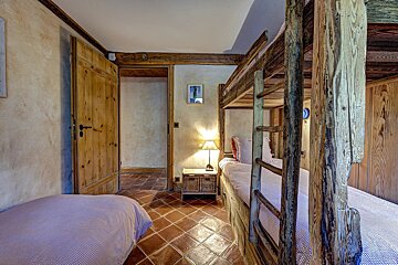 A rustic bedroom with sturdy wooden bunk beds, a single bed, and terracotta tiled floor. Exposed wooden beams and doors add to its cozy, charming feel.