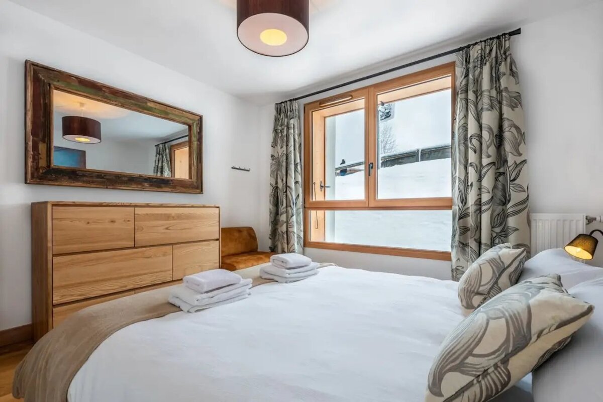 Cozy bedroom featuring a large bed, a wooden dresser and mirror, and a window looking out onto a snowy winter landscape.