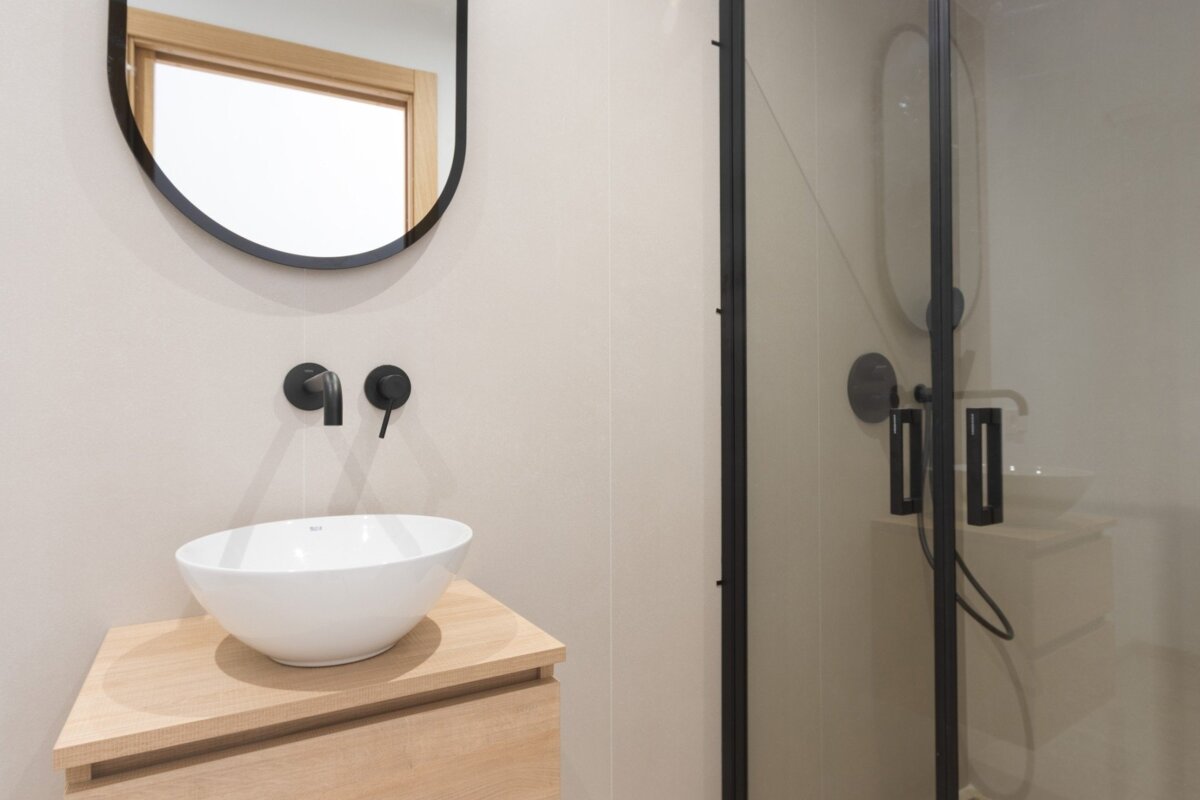 Modern bathroom features a black-framed mirror, wall-mounted black faucet over a white vessel sink on a wood vanity, next to a black-framed shower.